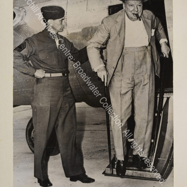 Photo shows WSC walking down the steps of a small aircraft. He is wearing a light suit and hat and has a cigar in his mouth.