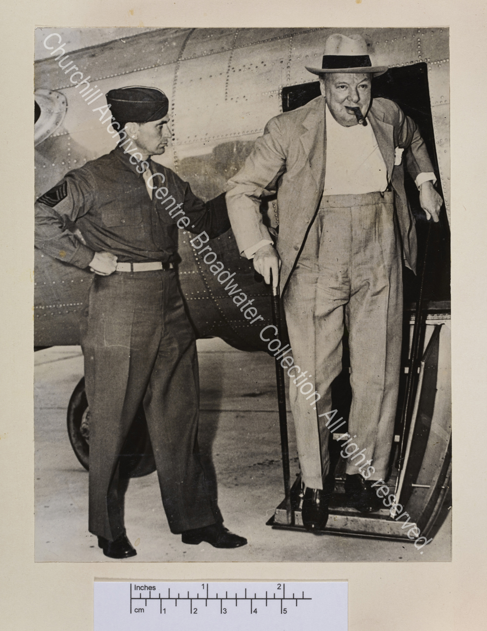 Photo shows WSC walking down the steps of a small aircraft. He is wearing a light suit and hat and has a cigar in his mouth.