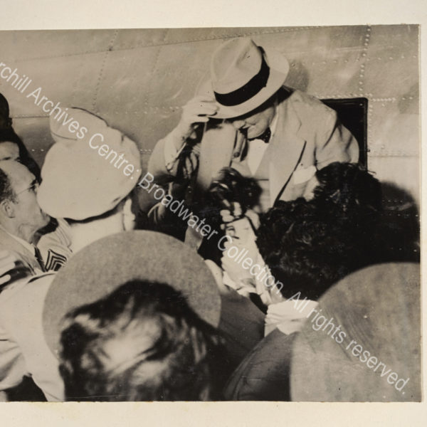 Photo shows WSC surrounded by a crowd of journalists as he disembarks from a small aircraft. He is wearing a light suit and hat and has a cigar in his mouth.