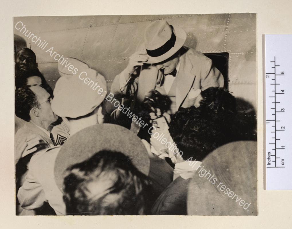 Photo shows WSC surrounded by a crowd of journalists as he disembarks from a small aircraft. He is wearing a light suit and hat and has a cigar in his mouth.