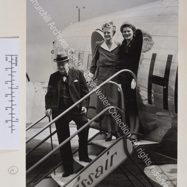 Photo shows WSC on the steps of a Swissair plane. He is wearing a pinstriped suit and waistcoat and a hat. CSC and Mary Churchill [later Lady Soames] stand together at the top of the steps smiling to the camera.