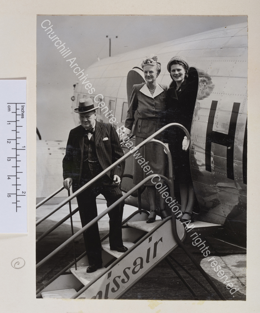 Photo shows WSC on the steps of a Swissair plane. He is wearing a pinstriped suit and waistcoat and a hat. CSC and Mary Churchill [later Lady Soames] stand together at the top of the steps smiling to the camera.