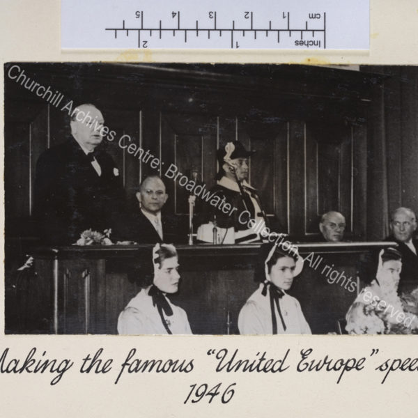 Photo shows WSC at the University of Zurich delivering his speech. Three young women wearing traditional bonnets sit in front of the lectern.