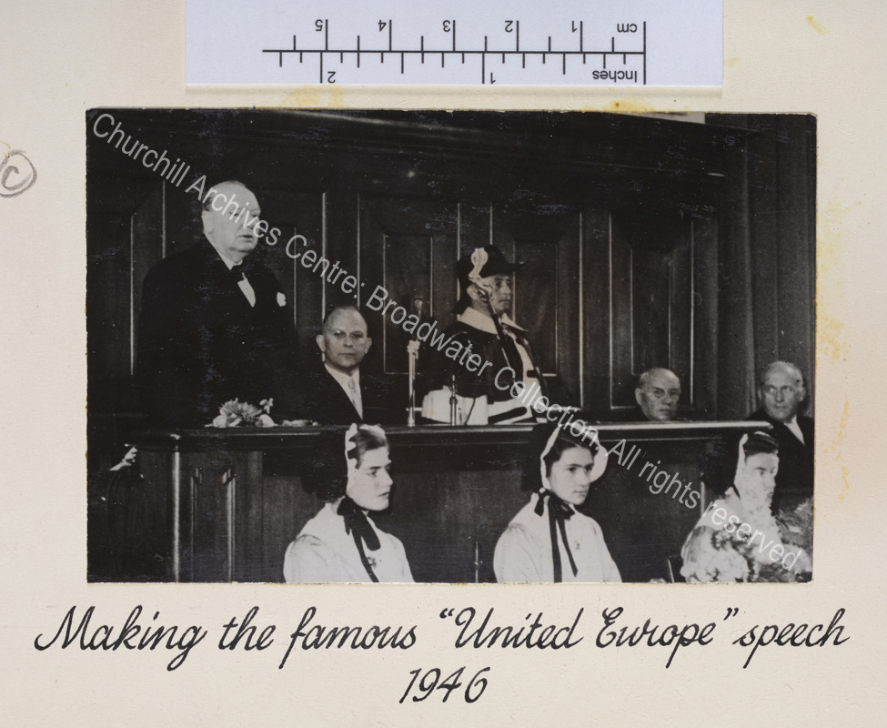 Photo shows WSC at the University of Zurich delivering his speech. Three young women wearing traditional bonnets sit in front of the lectern.