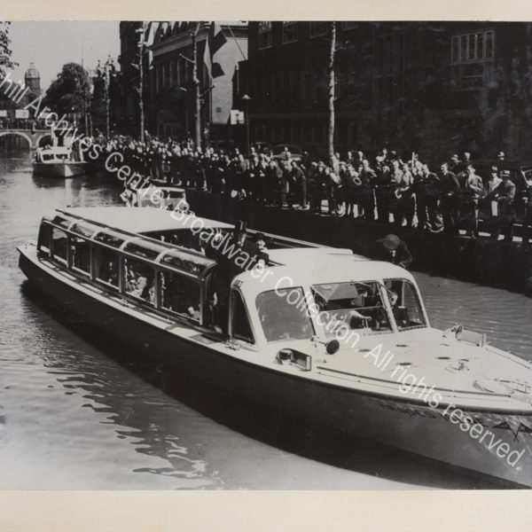 Photo shows WSC standing on board the Princess Irene wearing an overcoat and making a victory sign. Crowds of people are seen at the edge of the canal and standing on a bridge.