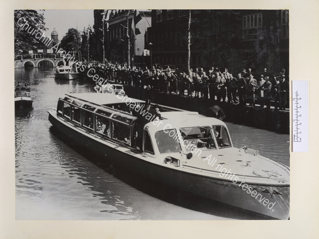 Photo shows WSC standing on board the Princess Irene wearing an overcoat and making a victory sign. Crowds of people are seen at the edge of the canal and standing on a bridge.
