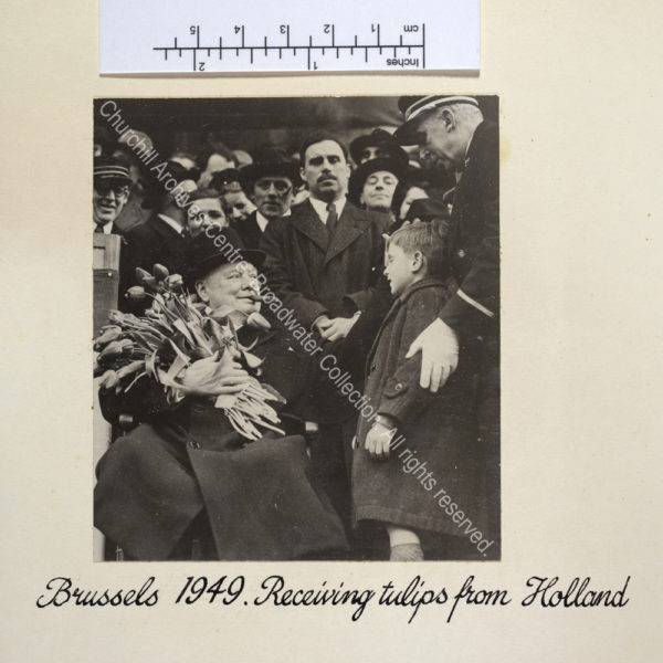 Photo shows WSC sitting in a chair with a cigar in his mouth and wearing an overcoat and hat. He is holding a large bunch of tulips which have been presented to him by a young boy standing to the right of the photo.