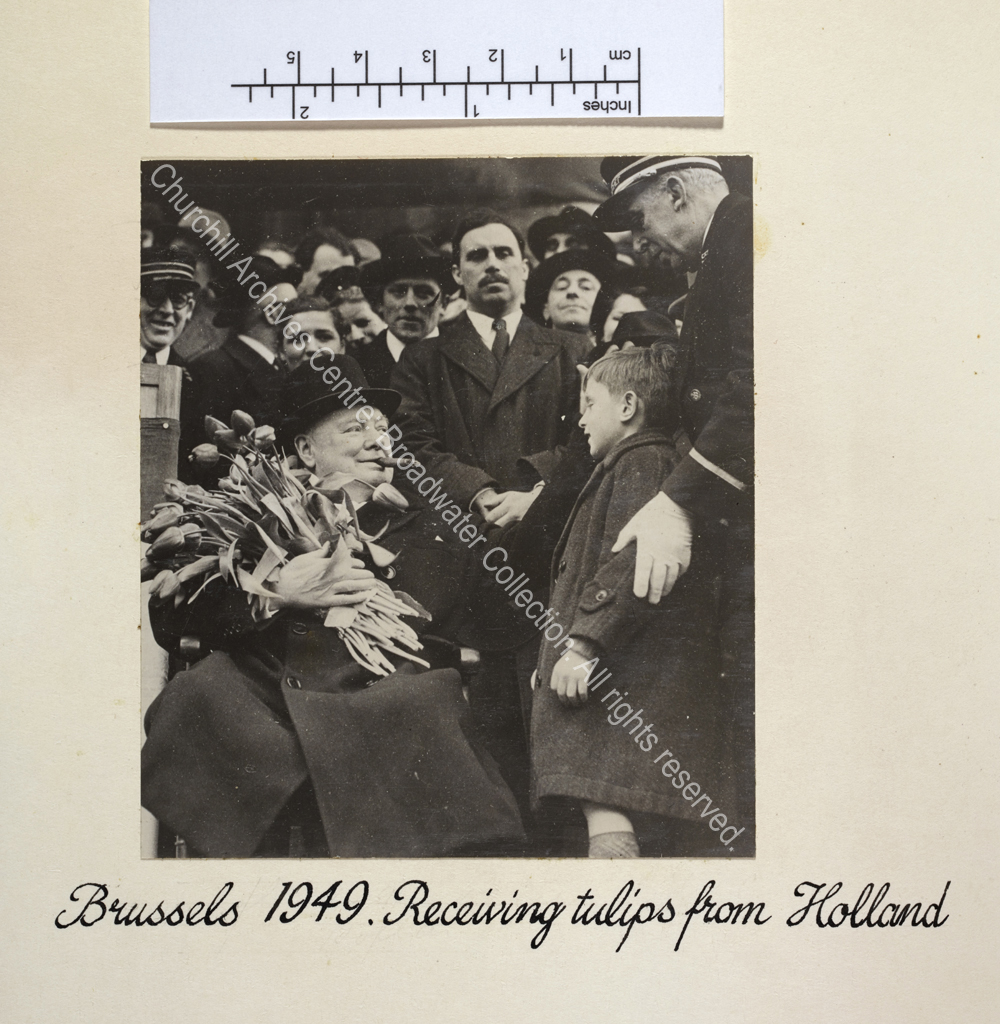 Photo shows WSC sitting in a chair with a cigar in his mouth and wearing an overcoat and hat. He is holding a large bunch of tulips which have been presented to him by a young boy standing to the right of the photo.