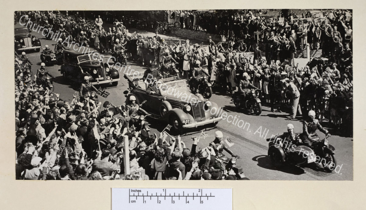 Photo taken from above showing WSC standing in an open topped car escorted by police on motorbikes. The sides of the street are crowded with people waving.