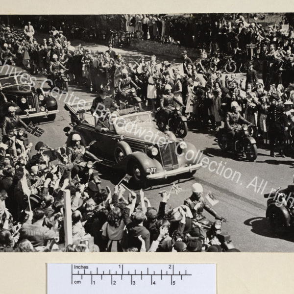 Photo taken from above showing WSC standing in an open topped car escorted by police on motorbikes. The sides of the street are crowded with people waving.