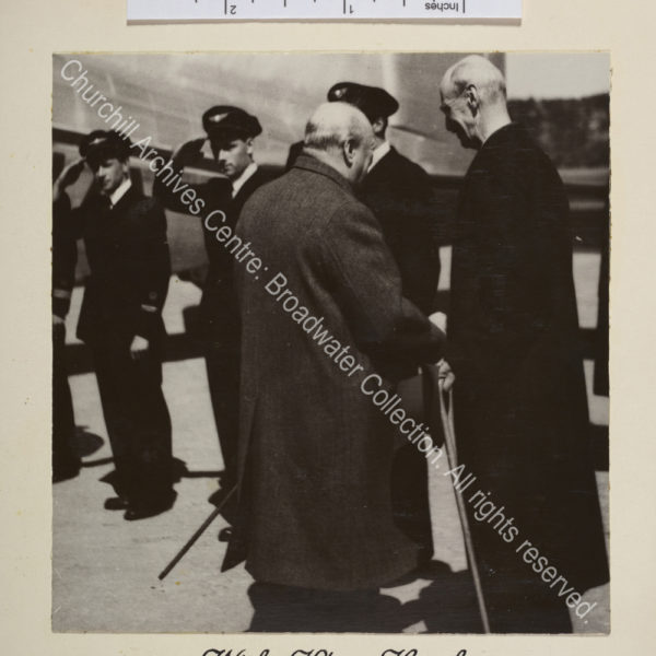 Photo shows WSC meeting King Haakon. Both are facing away from the camera. In the background there is an aeroplane and a line of men in uniform.
