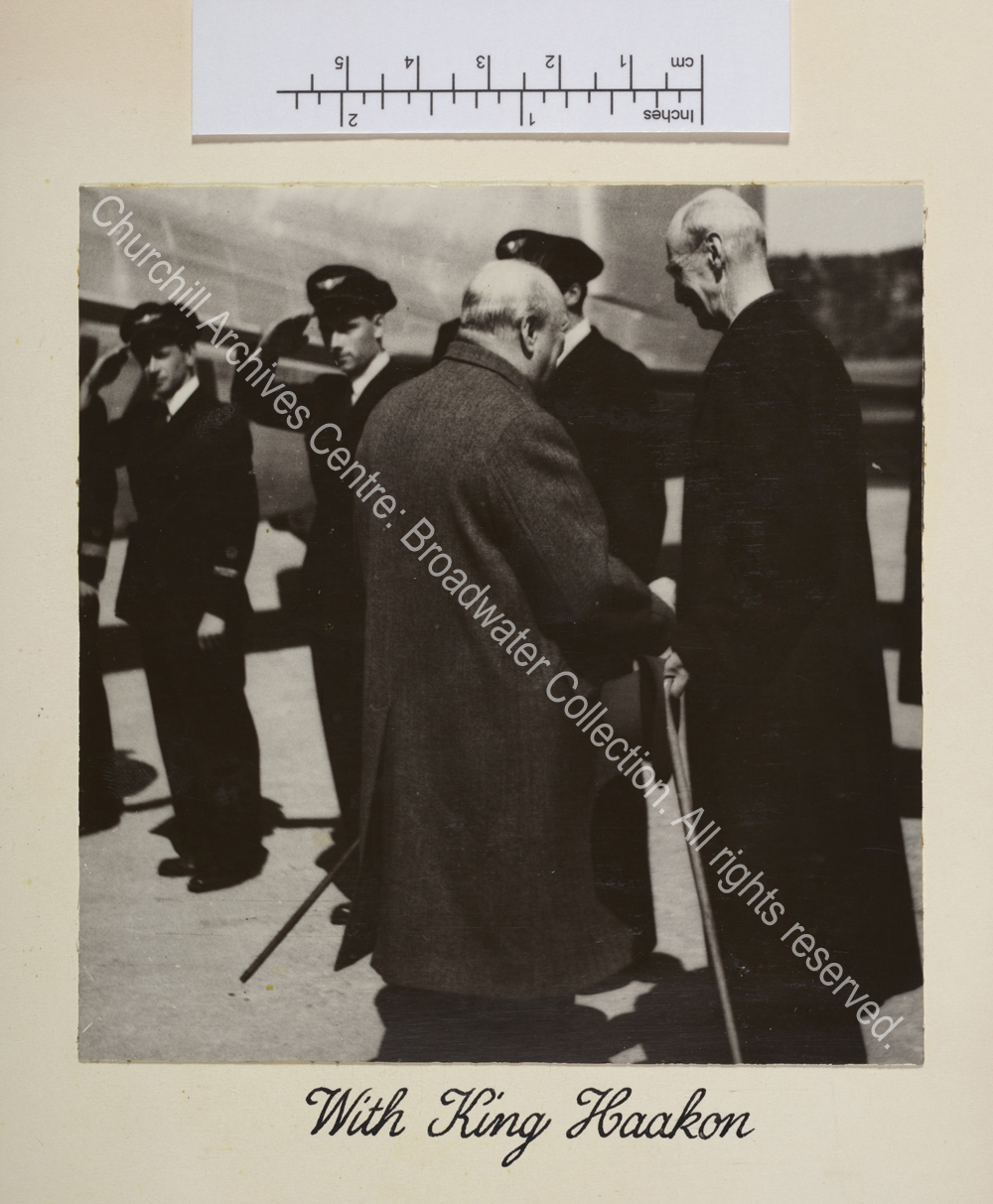 Photo shows WSC meeting King Haakon. Both are facing away from the camera. In the background there is an aeroplane and a line of men in uniform.