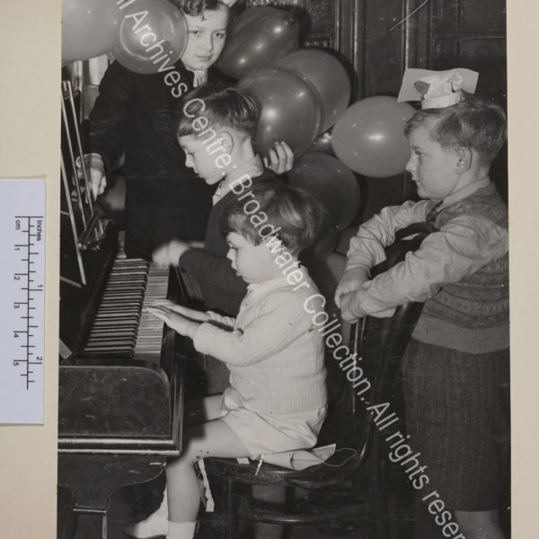 Photo of children at a piano. Some are wearing party hats and there are balloons in the background. Two boys are playing the piano [?Nicholas Soames is possibly the one in the foreground].