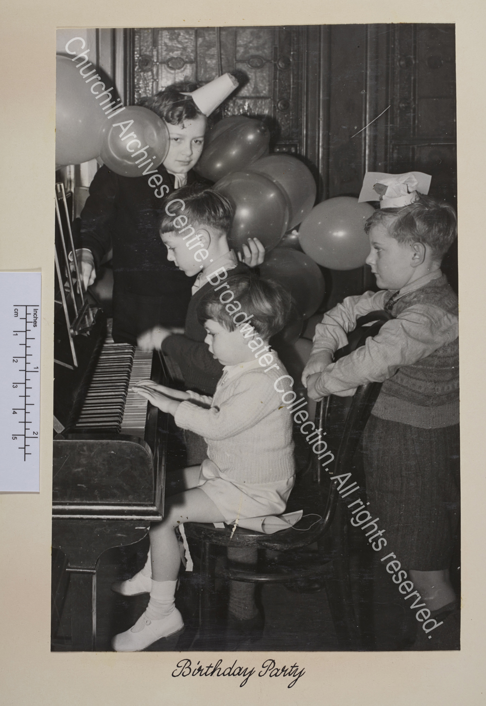 Photo of children at a piano. Some are wearing party hats and there are balloons in the background. Two boys are playing the piano [?Nicholas Soames is possibly the one in the foreground].