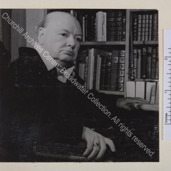 Head and shoulders portrait of WSC in half profile next to a bookshelf. WSC is wearing a siren suit and clasping the arm of his chair with one hand. He is keeping his place in a book with the other hand.