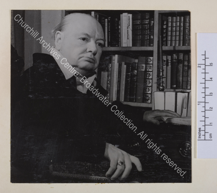 Head and shoulders portrait of WSC in half profile next to a bookshelf. WSC is wearing a siren suit and clasping the arm of his chair with one hand. He is keeping his place in a book with the other hand.