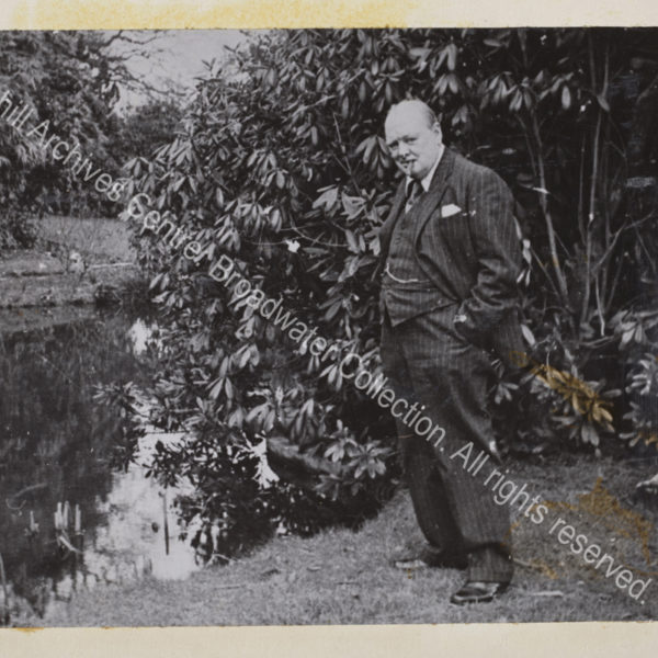 Photo of WSC standing next to a pond. He has a cigar in his mouth and his hands in his pockets and is wearing a pin-striped suit and waistcoat.