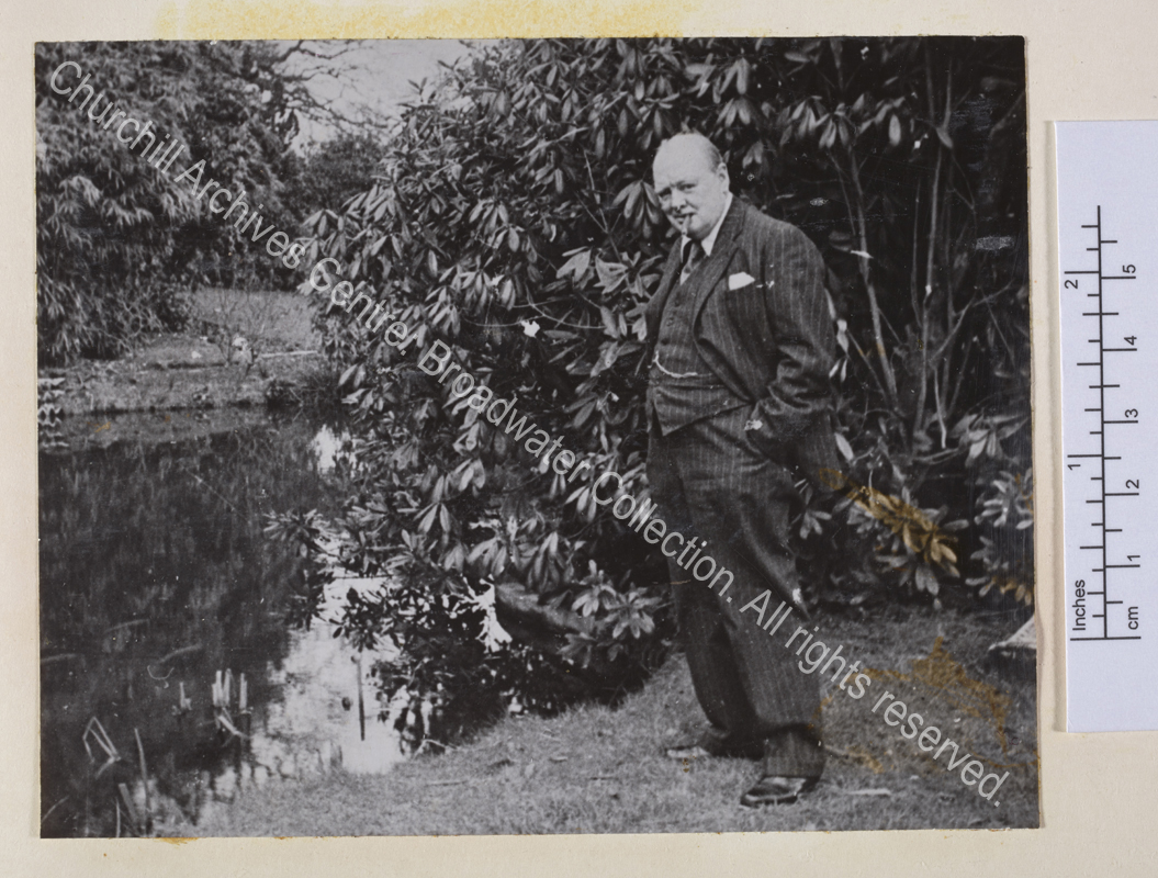 Photo of WSC standing next to a pond. He has a cigar in his mouth and his hands in his pockets and is wearing a pin-striped suit and waistcoat.