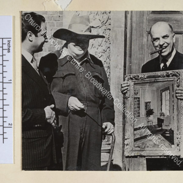 Photo shows WSC at the Chartwell entrance wearing a siren suit and a broad-brimmed sombrero hat with a feather in it. He is presenting his painting "The Blue Room