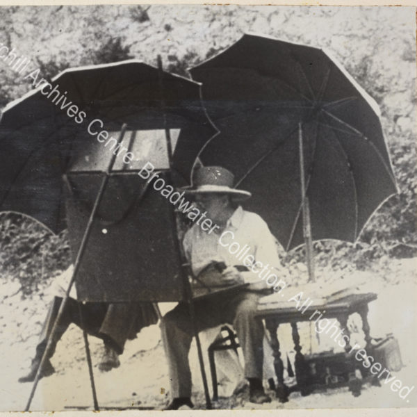 Photo shows WSC seated at a large easel absorbed in painting. He is wearing spectacles and a wide-brimmed hat and is shaded by two large umbrellas.