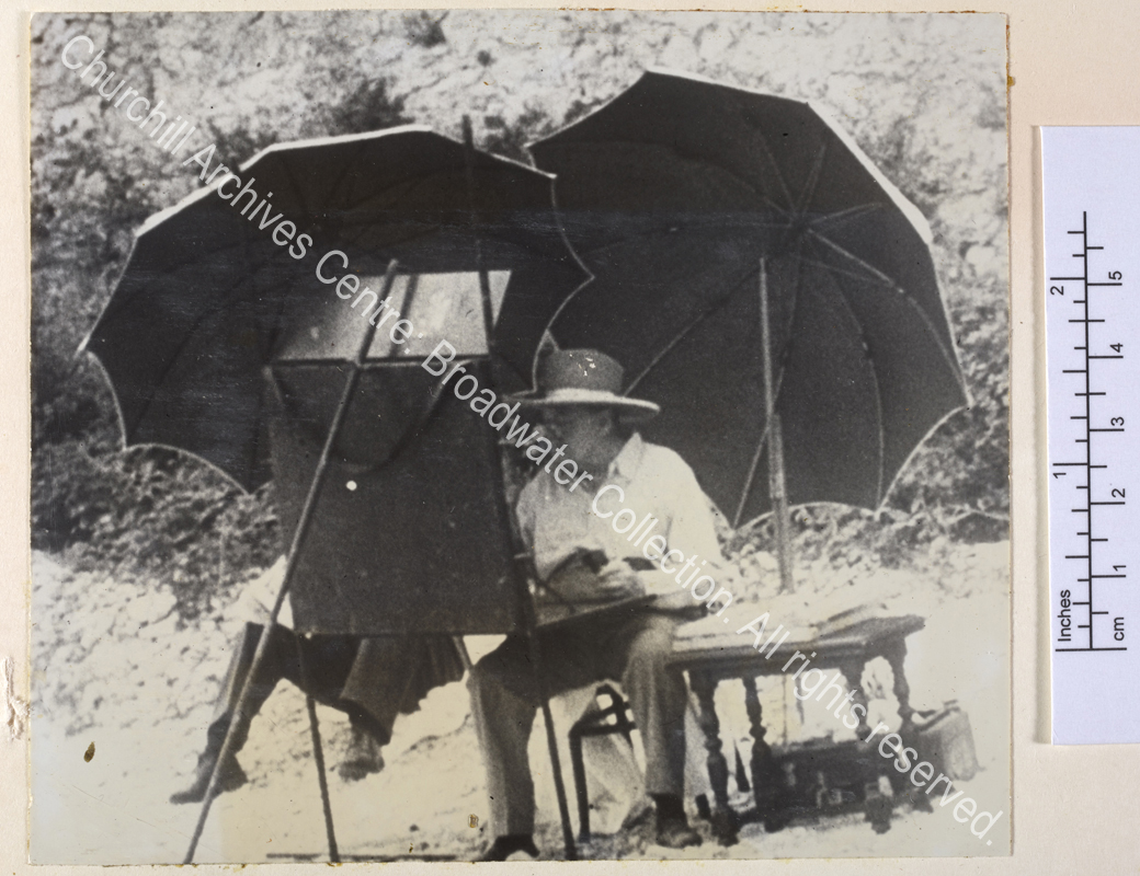 Photo shows WSC seated at a large easel absorbed in painting. He is wearing spectacles and a wide-brimmed hat and is shaded by two large umbrellas.