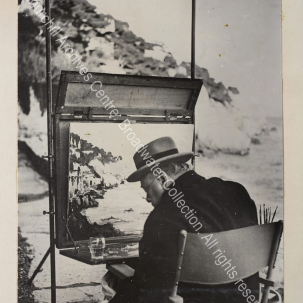 Photo taken from behind shows WSC painting near Cap d'Ail [France] with a glass resting on his painting table.