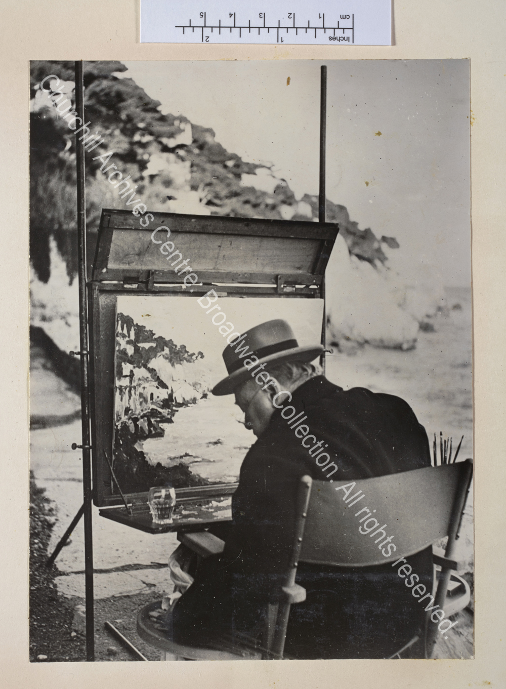 Photo taken from behind shows WSC painting near Cap d'Ail [France] with a glass resting on his painting table.