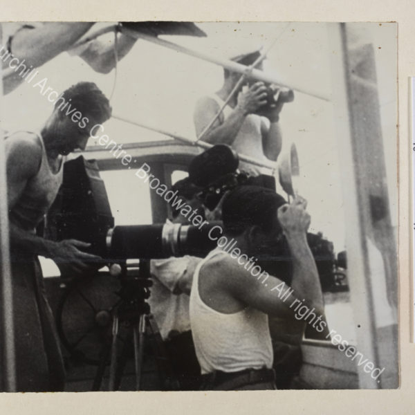 Photograph of a group of male press photographers in vests on board a boat. They are presumably photographing WSC while he paints.