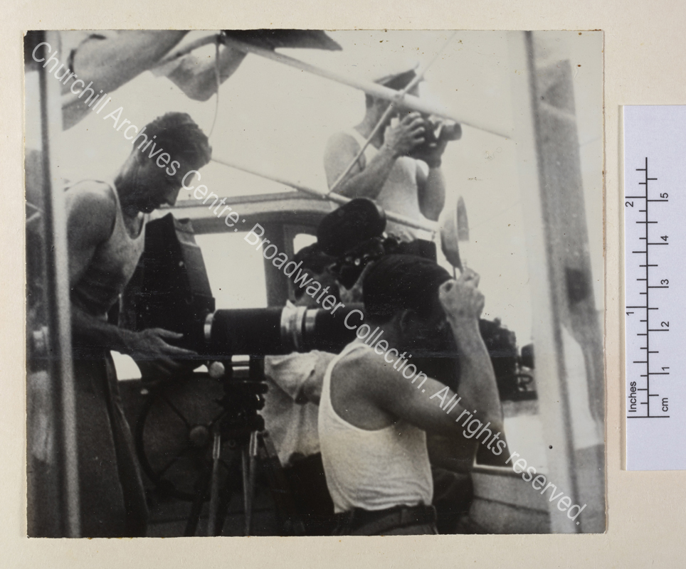 Photograph of a group of male press photographers in vests on board a boat. They are presumably photographing WSC while he paints.