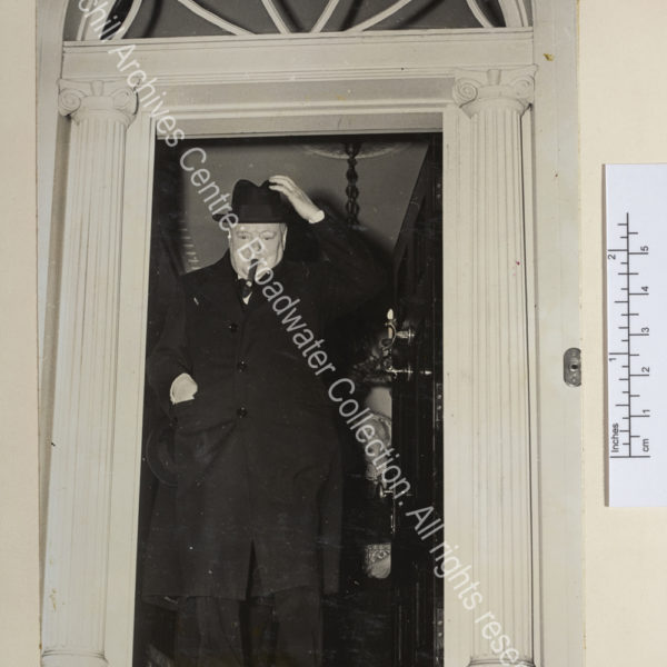 Photo shows WSC at the entrance of a grand property. He is wearing an overcoat and reaching for his hat. CSC is just seen smiling behind him.