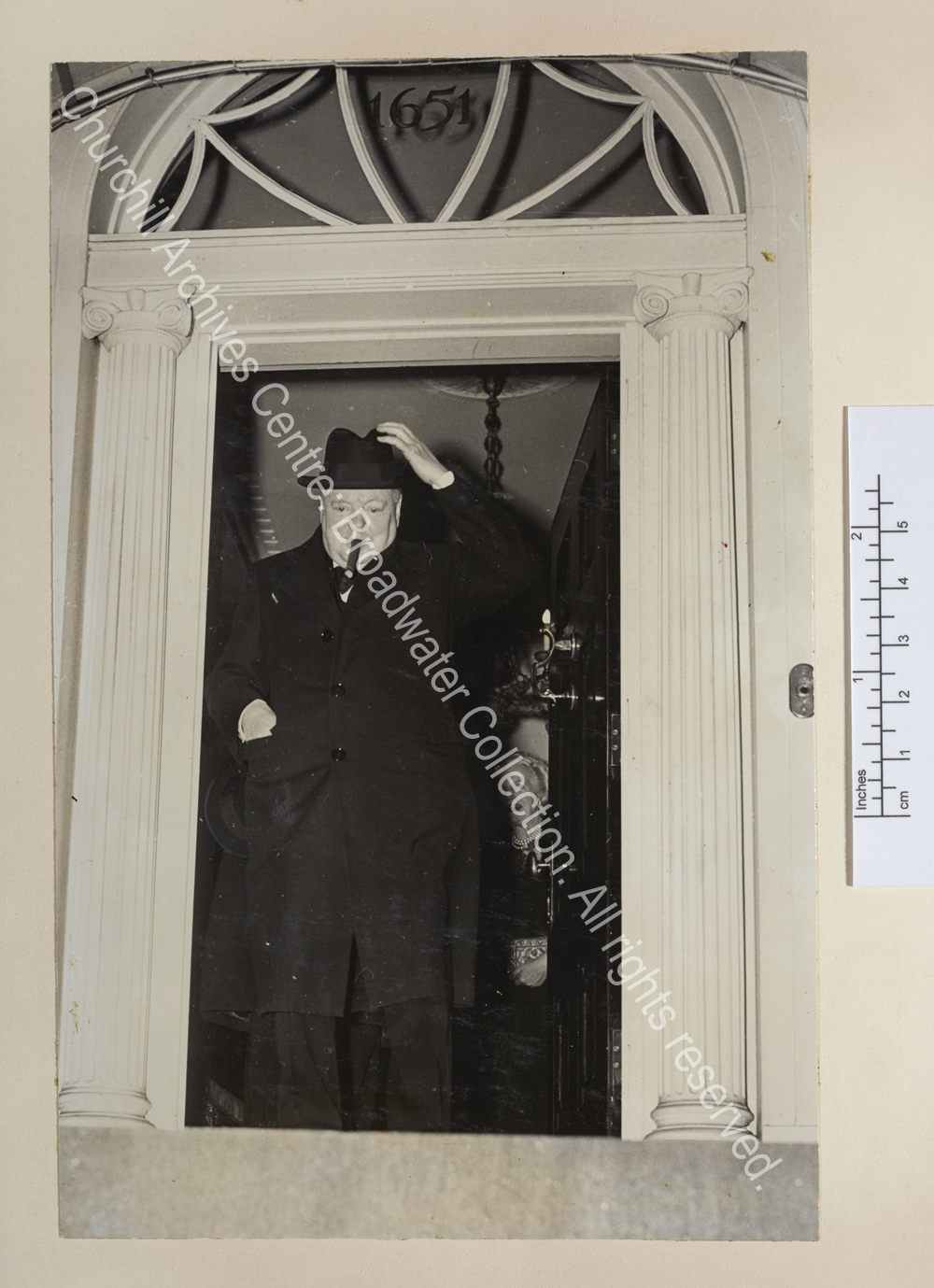 Photo shows WSC at the entrance of a grand property. He is wearing an overcoat and reaching for his hat. CSC is just seen smiling behind him.
