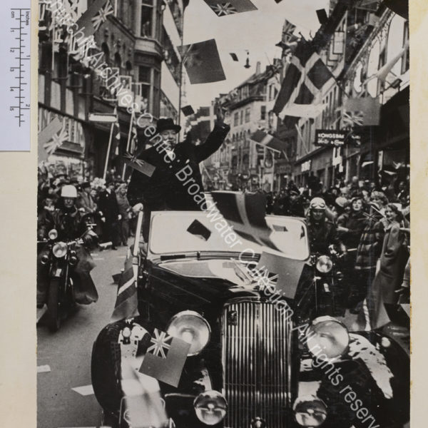 Photo shows WSC wearing an overcoat and hat standing in the back of an open topped car making the victory sign. The car is flanked by two motorcycle riders and the street is lined with crowds waving and cheering.