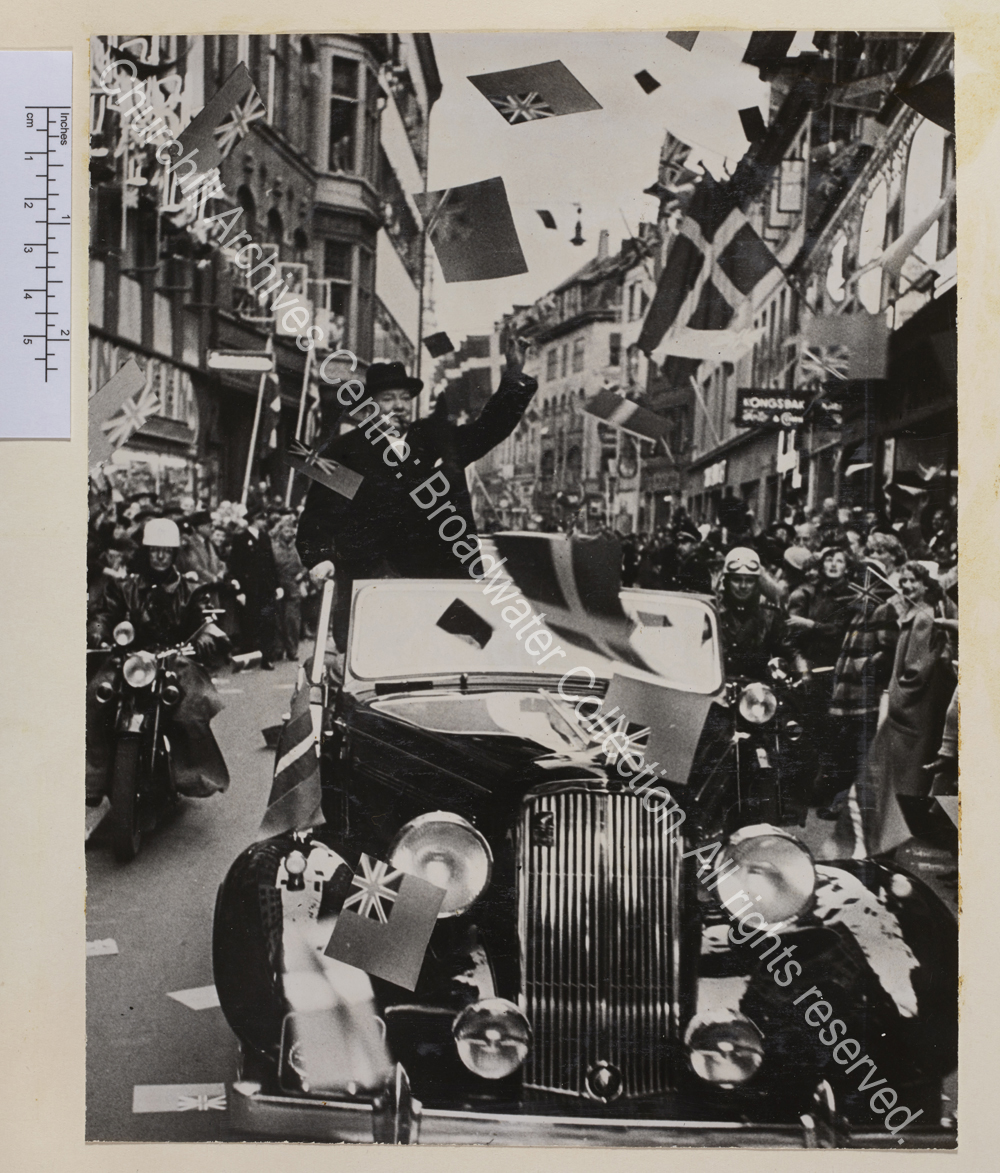 Photo shows WSC wearing an overcoat and hat standing in the back of an open topped car making the victory sign. The car is flanked by two motorcycle riders and the street is lined with crowds waving and cheering.