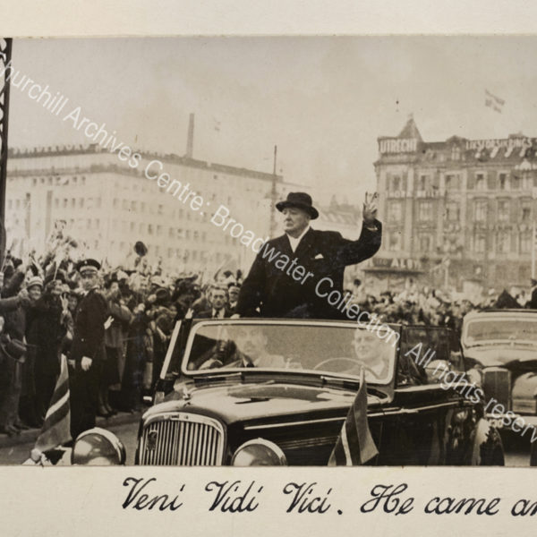Photo shows WSC wearing an overcoat and hat standing in the back of an open topped car making the victory sign. The street is lined with crowds waving and cheering.