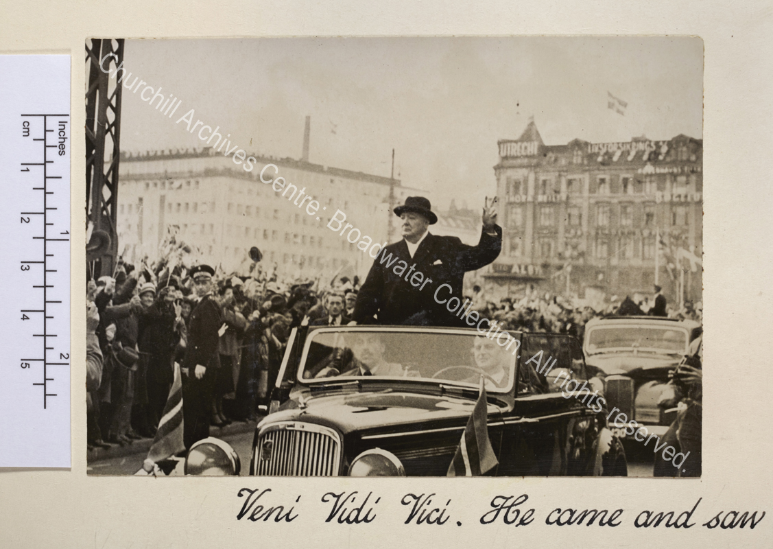 Photo shows WSC wearing an overcoat and hat standing in the back of an open topped car making the victory sign. The street is lined with crowds waving and cheering.