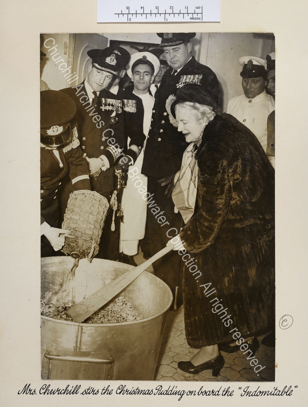 CSC is shown on board HMS Indomitable leaning over to use a large paddle to stir a huge pan. She is wearing a fur coat and is watched by naval officers and sailors.