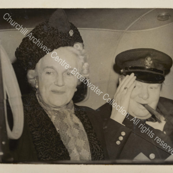 Photo shows WSC sitting together in the back of a car. WSC has a cigar in his mouth and raises his hand to his peaked cap.