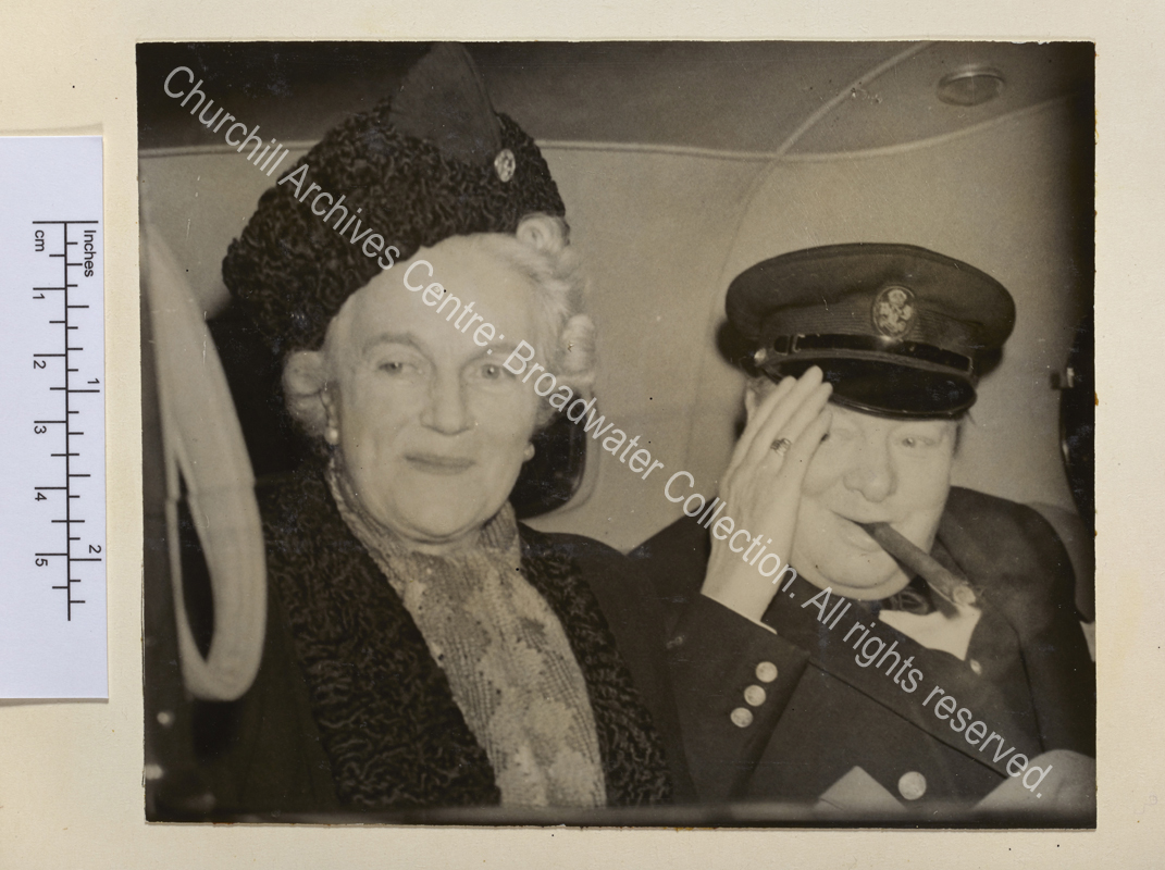 Photo shows WSC sitting together in the back of a car. WSC has a cigar in his mouth and raises his hand to his peaked cap.