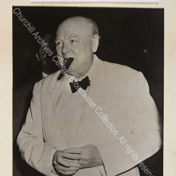 Photo shows WSC standing with a cigar in his mouth. He is wearing a white jacket and black bow tie and is fingering the ring on his finger.