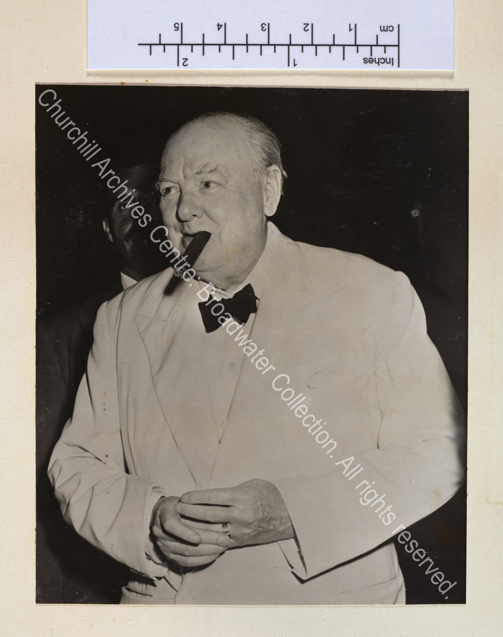 Photo shows WSC standing with a cigar in his mouth. He is wearing a white jacket and black bow tie and is fingering the ring on his finger.