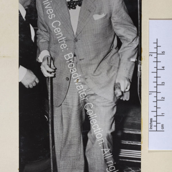 Photo shows WSC wearing a pale coloured suit and hat stepping out of a car. He has a cigar in his mouth and is smiling.