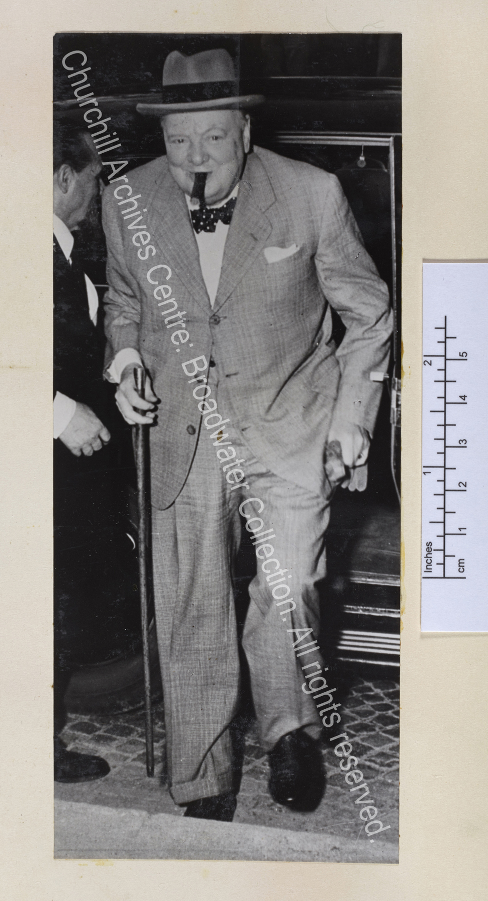Photo shows WSC wearing a pale coloured suit and hat stepping out of a car. He has a cigar in his mouth and is smiling.