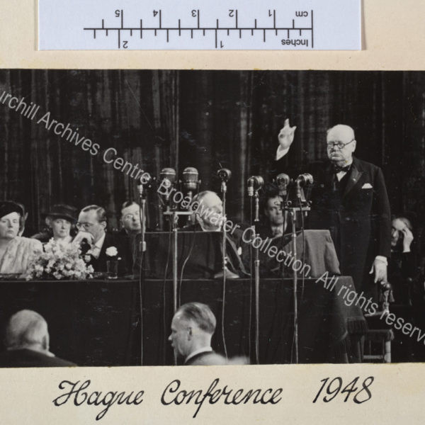 Photo of WSC speaking in front of a bank of microphones with his arm raised high.