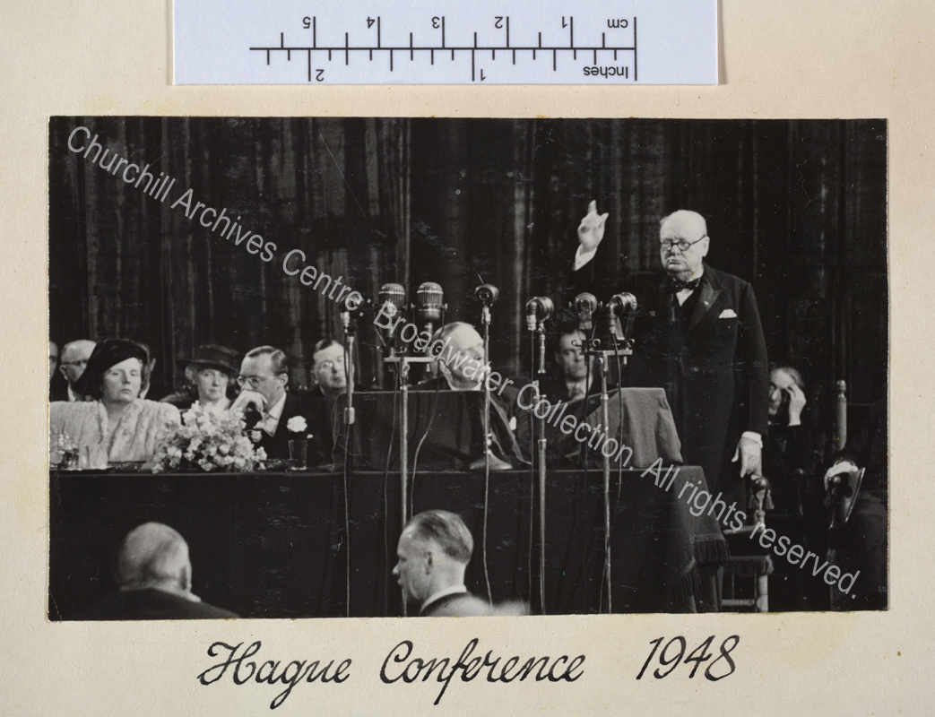 Photo of WSC speaking in front of a bank of microphones with his arm raised high.