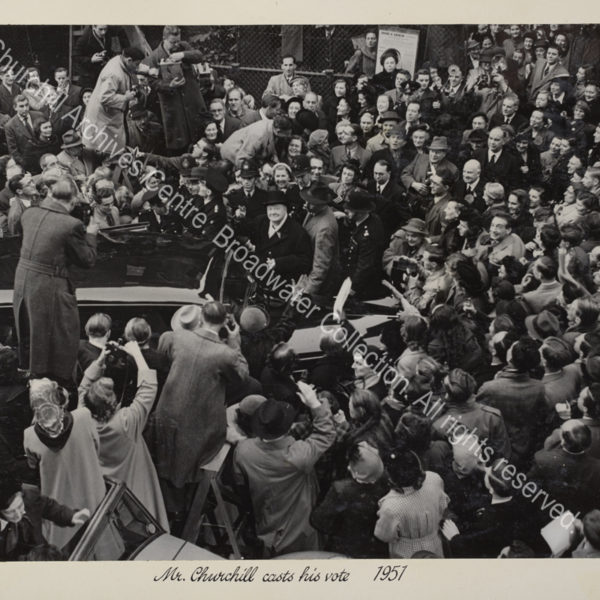 Photo of WSC at the 1951 General Election. The photo is taken from a high vantage point and shows WSC next to a car and surrounded by a ring of policemen and a dense crowd. WSC is smiling and looking up at the camera as he raises his hand to give a victory salute.