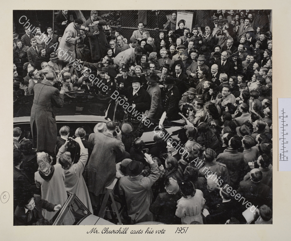 Photo of WSC at the 1951 General Election. The photo is taken from a high vantage point and shows WSC next to a car and surrounded by a ring of policemen and a dense crowd. WSC is smiling and looking up at the camera as he raises his hand to give a victory salute.