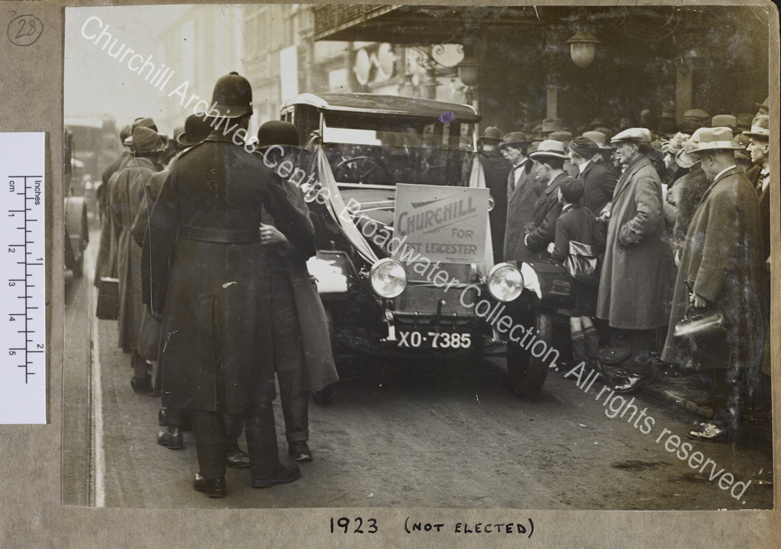 Photograph of a Wolseley car (registration XO 7385) used during the General Election for WSC's unsuccessful campaign to return to Parliament as MP for West Leicester.  The car has the sign "Churchill for West Leicester" on the front and is driving along a street with crowds of people on either side looking in [presumably with WSC inside