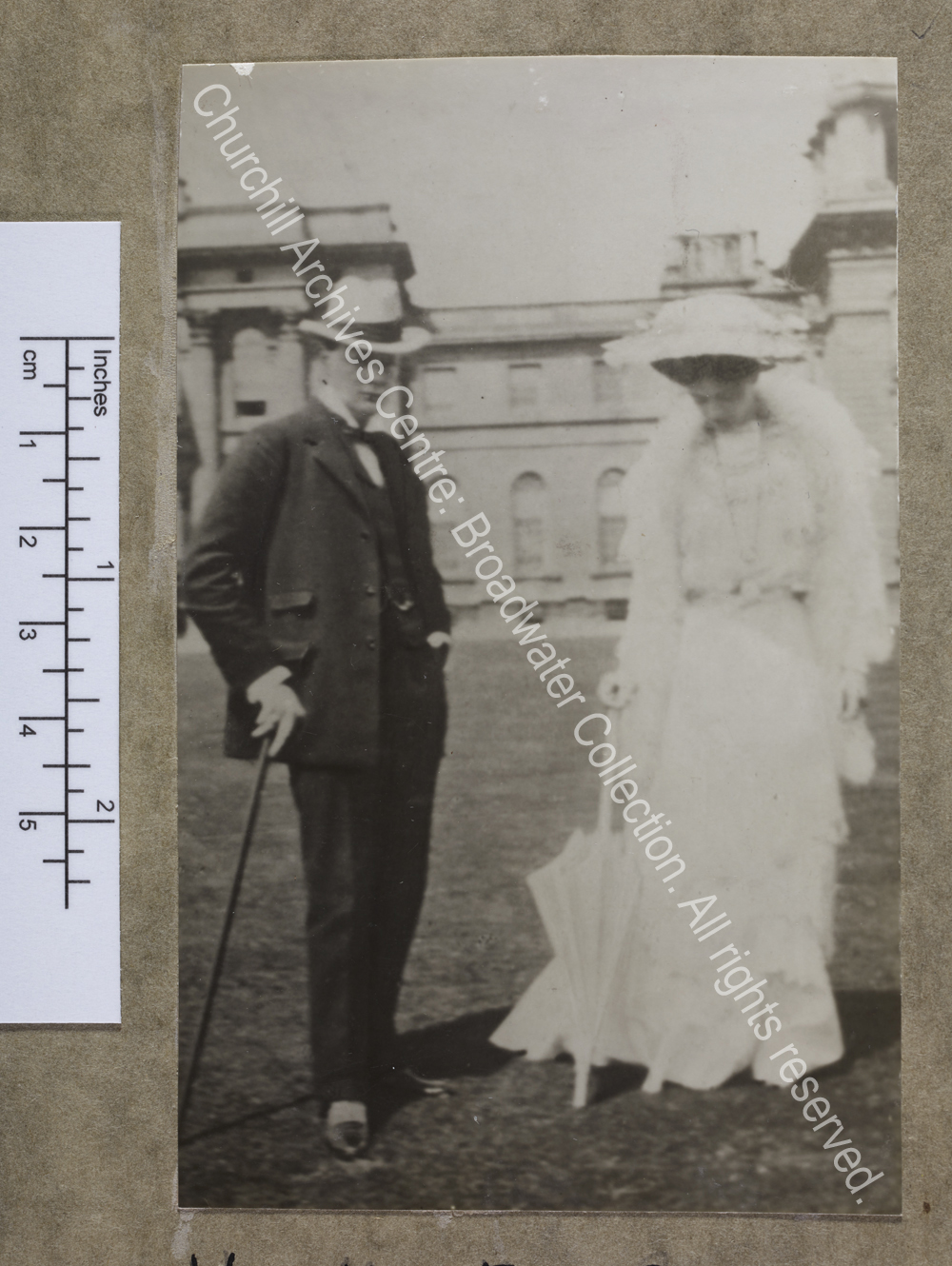 Full-length photograph of WSC with Ethel Barrymore standing on the lawn outside Blenheim Palace [Oxfordshire].  Ethel Barrymore is wearing a long white dress with a white feather boa and a large hat