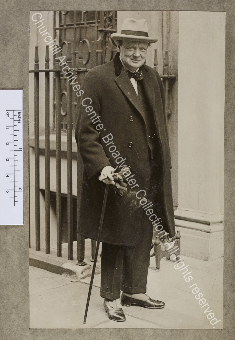 Full-length photograph of WSC [Chancellor of the Exchequer] standing on the pavement by the railings of 11 Downing Street [London].  He is smiling at the camera