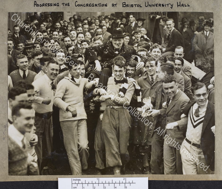Photograph of WSC being carried on the shoulders of Bristol University students through a crowd [to the Students' Union].  He is wearing a dark overcoat and homburg hat